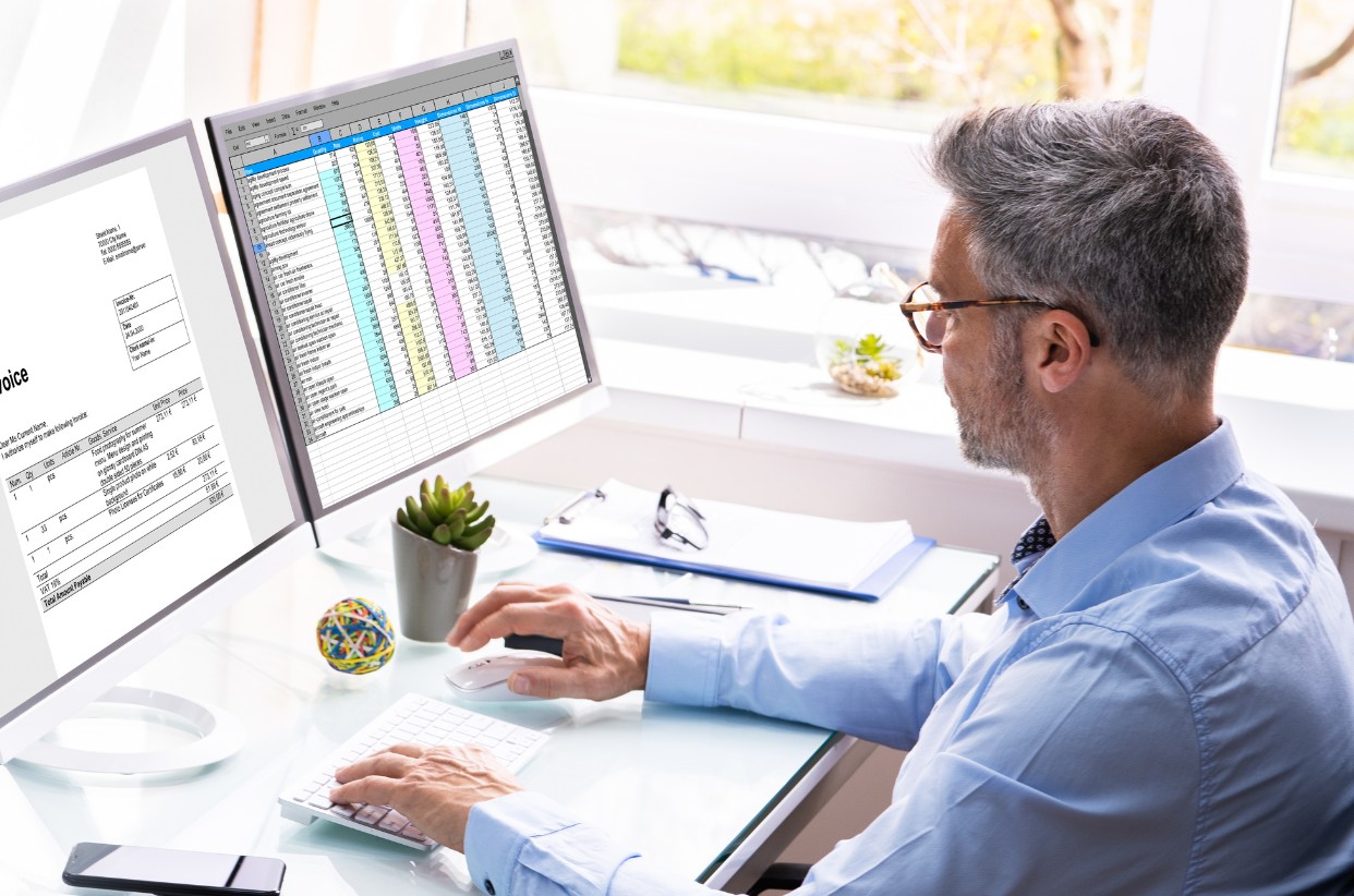Un professionnel en bureau travaillant sur un double écran affichant des tableaux de données et des factures, illustrant l'interface de gestion de la solution HABIGED